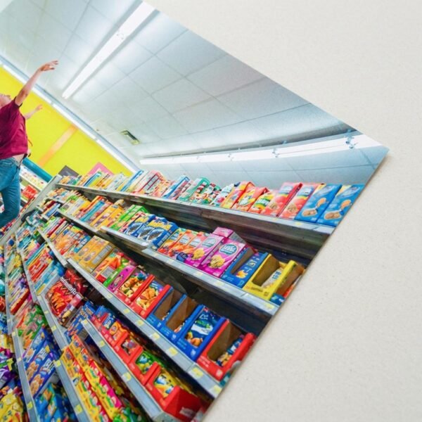 Photographie d'une figure humaine en suspension horizontale au centre d'un environnement commercial symétrique. L'image se concentre sur la désarticulation des membres et la saturation chromatique des produits de grande consommation.